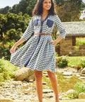 A woman standing outdoors in a casual chic setting, wearing a blue and white checkered shirtdress with long sleeves, a round neckline, and a belt.